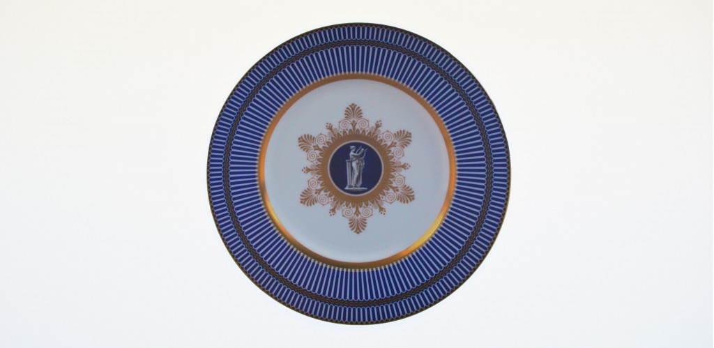 A ceramic blue, gold and white plate positioned in the center of the image, placed on a clear white wall background at world of wedgwood