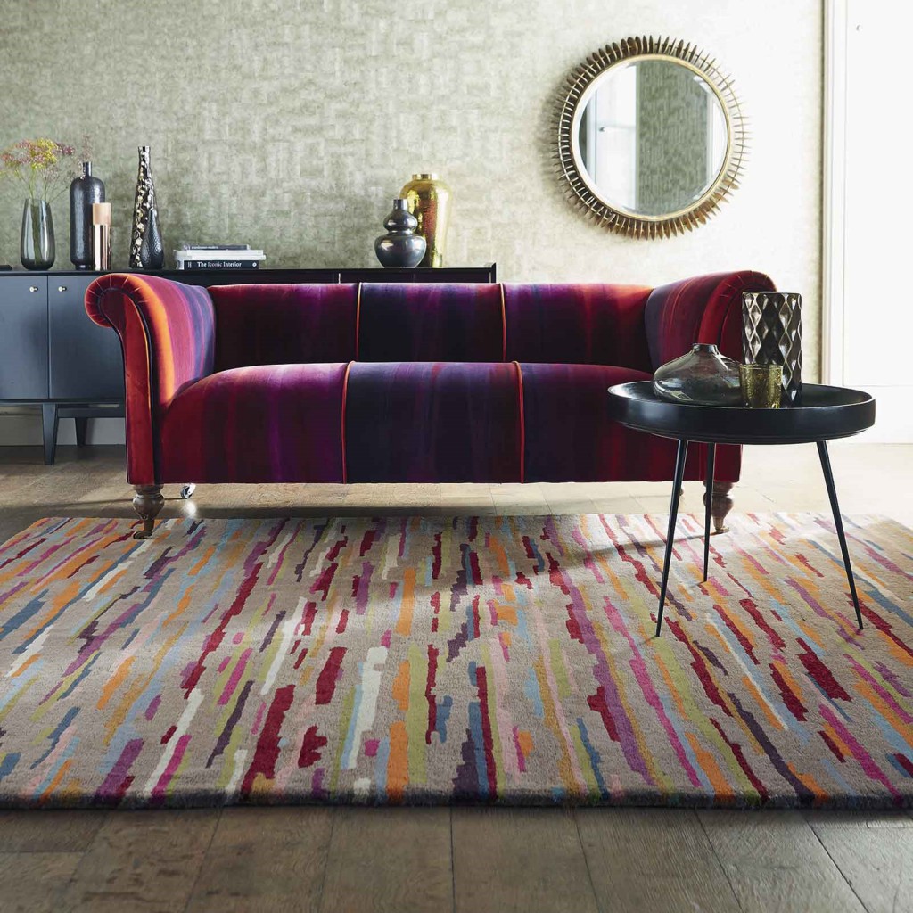 a bright purple, red and orange sofa sits underneath a multicoloured striped rug