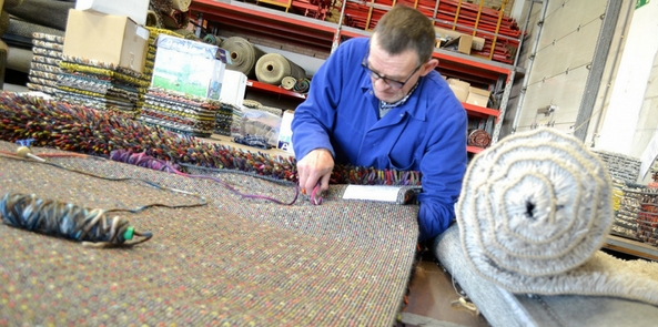 a man showing how a rug is made while weaving a rug from the back using a tool in a factory setting